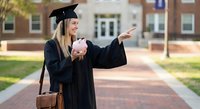 Young blonde woman graduate in black cap and gown pointing forward on university campus pathway, celebrating academic achievement and future opportunities.