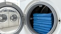 Stack of blue towels neatly folded inside open white washing machine drum in modern laundry room with additional machines visible in background.