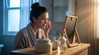 Woman with dark hair and medium skin tone applying skincare product to face while looking in mirror during morning beauty routine in bright bathroom setting.