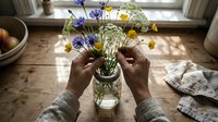 Person with dark skin tone arranging fresh wildflowers in glass vase on wooden table near window, hands holding stems of blue and yellow blooms with greenery for home decoration and floral design.
