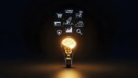 Glowing light bulb with brain filament and business icons floating above on dark background representing innovation and creative thinking in commerce.
