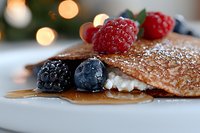 Fresh crepes filled with cream, topped with ripe raspberries, blackberries and blueberries, dusted with powdered sugar and drizzled with maple syrup on white plate.