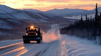 Snow plow truck clearing winter road at sunset with mountain silhouettes and evergreen forest, creating dramatic light trails in snowy conditions.