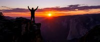 Male hiker celebrating success on mountain cliff edge during golden hour sunrise with arms raised in victory pose overlooking dramatic canyon landscape.