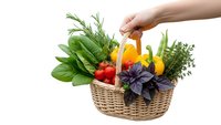 Hand holding wicker basket filled with fresh organic herbs and vegetables including basil, rosemary, thyme, peppers on white background isolated.