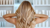 Blonde woman with long wavy hair shown from behind in hair salon. Professional hairstyle with natural highlights and soft waves demonstrates modern balayage technique.