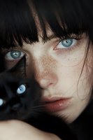 Close-up portrait of young woman with striking blue eyes, dark bangs, and freckles alongside black cat with matching blue eyes creating mysterious atmosphere.