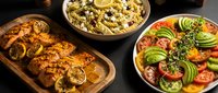 Grilled chicken with lemon slices, creamy pasta salad and fresh avocado tomato salad arranged on wooden table for healthy meal planning and cooking.