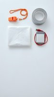 Emergency survival kit flat lay with orange whistle, duct tape, power bank, and rain poncho on white background, top view.