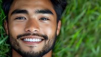 Young Asian man with beard smiling outdoors against green foliage background. Happy male portrait with natural lighting for lifestyle and wellness content.