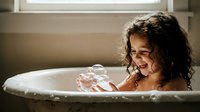 Happy young girl with curly hair enjoying bath time, playing with bubbles in white bathtub. Childhood hygiene and self-care routine concept.