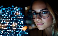 Young Caucasian woman wearing glasses illuminated by blue bokeh lights at night, close up portrait with soft dreamy expression and glowing city backdrop.