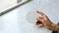 Female hand with medium skin tone holding blank white circular disc on marble surface, suitable for product mockups and branding presentations.
