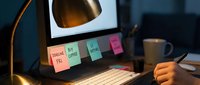 Workspace organization with sticky notes on monitor, desk lamp, keyboard, and coffee mug. Productivity and planning setup for remote work or office environment.