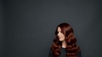 Woman with long, voluminous wavy auburn hair styled in loose waves, looking to the side with a subtle smile against a neutral dark gray background. Suitable for beauty, hair care, and salon marketing materials.