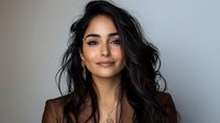 Young woman with long dark wavy hair and natural makeup wearing brown blazer smiling confidently against gray background, professional headshot style portrait.
