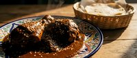 Tender chicken smothered in rich dark mole sauce with sesame seeds on a decorative ceramic plate, warm sunlight, rustic wooden table.