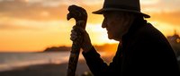 Elderly man with carved walking stick silhouetted against golden sunset on beach. Senior gentleman enjoying peaceful evening by ocean waves during golden hour.