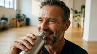 Middle-aged man with full beard and dark hair drinking water from a bottle indoors, wellness and hydration concept.