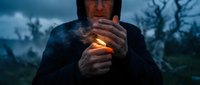 Caucasian man in black hoodie lighting a flame outdoors at dusk, moody dark atmosphere with bare trees and dramatic cloudy sky.