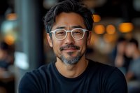Confident mature Asian man with glasses and salt-pepper beard smiling warmly at camera. Professional portrait with soft bokeh background in casual business setting.