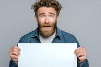 Surprised Caucasian man with curly hair and full beard holding blank white poster against gray background, perfect for adding custom message or advertisement.