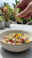 Hand squeezing fresh lime juice over colorful ceviche salad with diced fish, corn, red onion, and cilantro in ceramic bowl outdoors.
