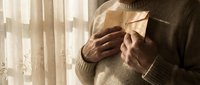 Mature Caucasian man in beige sweater holding paper airplane by sunny window with warm natural lighting creating peaceful contemplative mood.