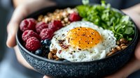 Healthy breakfast bowl with sunny side up egg, fresh berries, granola, and greens served in dark ceramic bowl. Modern food styling for restaurant menu and blogs.