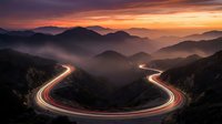 Winding mountain road with light trails at sunset creating dramatic curves through misty valley landscape with layered hills and golden sky.