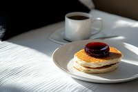 Stack of fluffy pancakes topped with berry jam on white plate next to coffee cup in morning sunlight creating dramatic shadows on textured tablecloth.