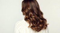 Young woman with long wavy brown hair wearing white knit sweater from behind showing healthy shiny hair texture and natural highlights for beauty salon.