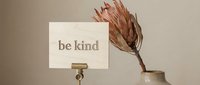 Wooden sign with engraved be kind message in a metal holder beside a dried protea flower in a ceramic vase, warm neutral tones.