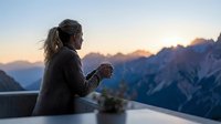 Woman with long dark hair wearing casual athletic jacket sitting on railing overlooking mountain valley at sunrise, contemplative outdoor lifestyle moment for travel and wellness content.