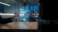 Businessman hand touching holographic data analytics charts and graphs floating above wooden desk in modern office environment with blurred team background.