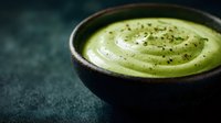Creamy green matcha latte in rustic ceramic bowl with foam art and black sesame seeds on dark textured background.