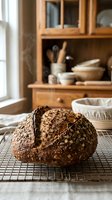 Freshly baked artisan multigrain bread loaf cooling on wire rack in rustic farmhouse kitchen with wooden hutch displaying ceramic bowls and utensils.