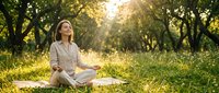 Caucasian woman in light casual clothing meditating cross-legged on grass under tree canopy during golden hour, practicing mindfulness and wellness outdoors.