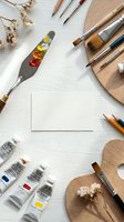 Artist workspace flatlay with paint tubes, brushes, palette knives, wooden palettes, and blank card on white wood, top view.