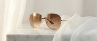 Stylish aviator sunglasses with golden frames and gradient brown lenses resting on white fabric surface in soft natural lighting for fashion accessories.
