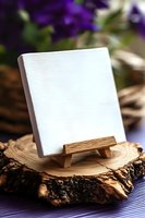 Blank white canvas on wooden mini easel displayed on rustic wood slice with purple floral background, creating artistic composition for mockup presentation.