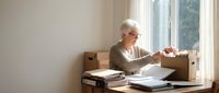 Senior Caucasian woman with white hair and glasses unpacking cardboard box at wooden desk near window in bright room during relocation or moving day.