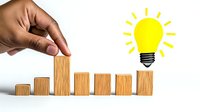 African American hand arranging wooden blocks in ascending order with bright yellow light bulb icon, representing growth, innovation and business success concept on white background.