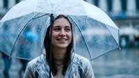 Young woman with long dark hair and fair complexion holding transparent umbrella in rainy weather, wearing knit sweater, smiling outdoors.
