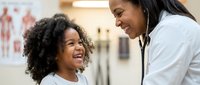 African American female pediatrician examining happy young Black girl patient with stethoscope in modern medical clinic office during routine checkup visit.