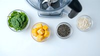 Healthy smoothie ingredients with spinach, mango, chia seeds and protein powder arranged around blender for nutritious breakfast preparation.