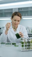 Female scientist of European descent examining green plant samples in laboratory setting with test tubes and specimens for research and biotechnology applications.