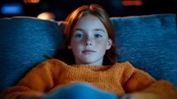 Young redhead girl in orange knit sweater relaxing on blue couch, illuminated by warm ambient lighting creating cozy atmospheric mood for lifestyle and interior design concepts.