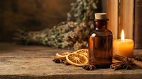 Amber glass bottle with cork stopper surrounded by dried orange slices, cinnamon sticks, star anise and burning candle on rustic wooden table for aromatherapy.