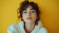 Young woman with curly brown hair wearing white hoodie against bright yellow background. Casual portrait with natural makeup and confident expression.